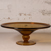 An amber brown translucent glass pedestal plate with a flared rim and a decorative stem base, set against a textured off-white wall.