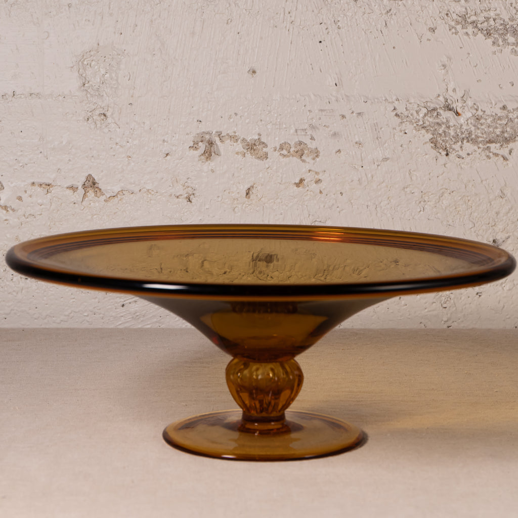 An amber brown translucent glass pedestal plate with a flared rim and a decorative stem base, set against a textured off-white wall.