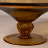 Close-up of an amber glass pedestal bowl or candle holder with a flared round base and a faceted stem, photographed on a neutral light beige surface.