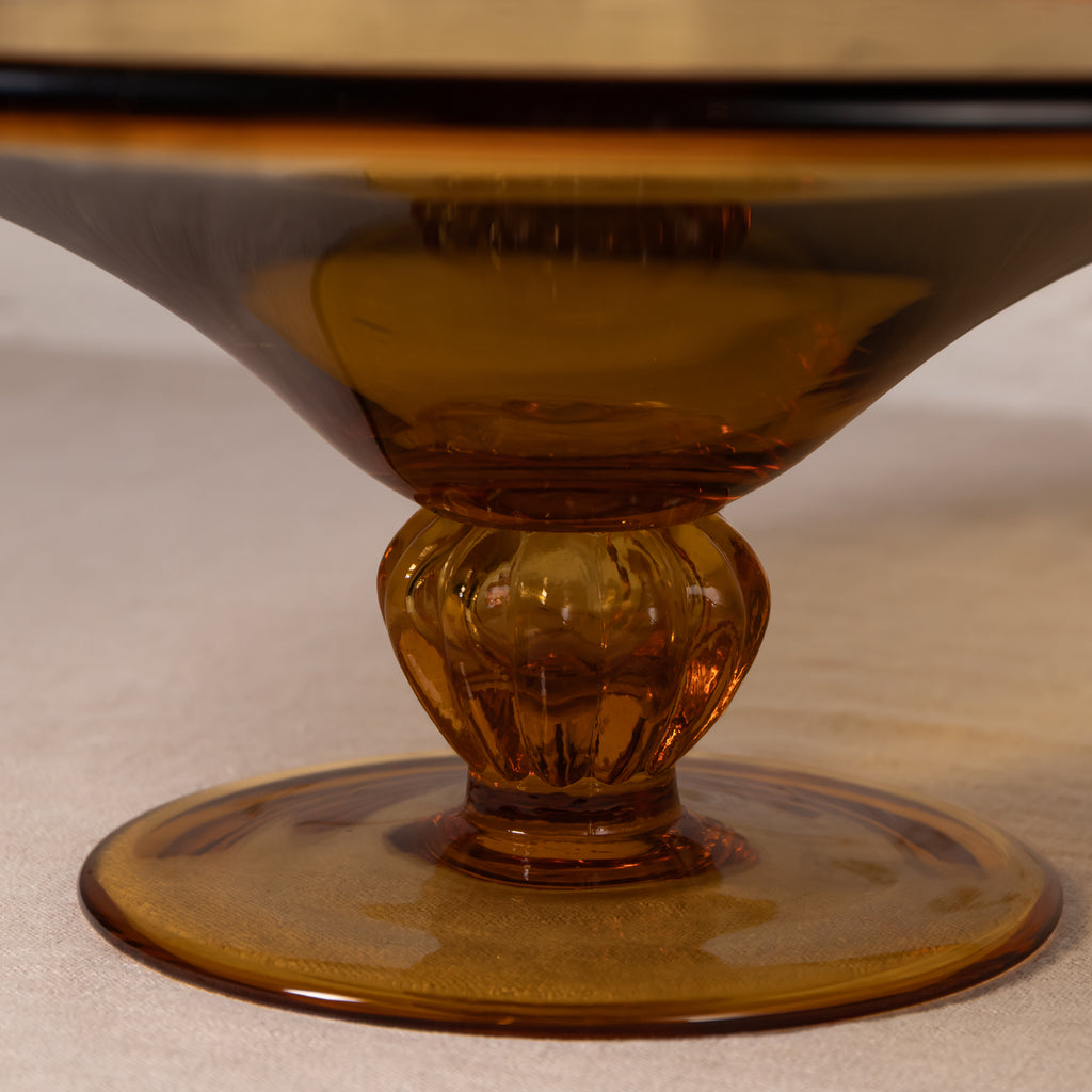 Close-up of an amber glass pedestal bowl or candle holder with a flared round base and a faceted stem, photographed on a neutral light beige surface.