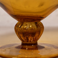 Close-up of an amber glass goblet or cup with a flared rim, ribbed stem, and circular base, photographed against a softly blurred neutral background.