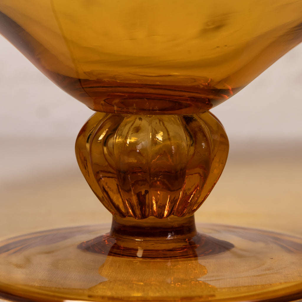 Close-up of an amber glass goblet or cup with a flared rim, ribbed stem, and circular base, photographed against a softly blurred neutral background.