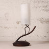 A table lamp with a frosted white cylindrical glass shade and a dark brown sculptural metal base, set against a textured off-white wall.
