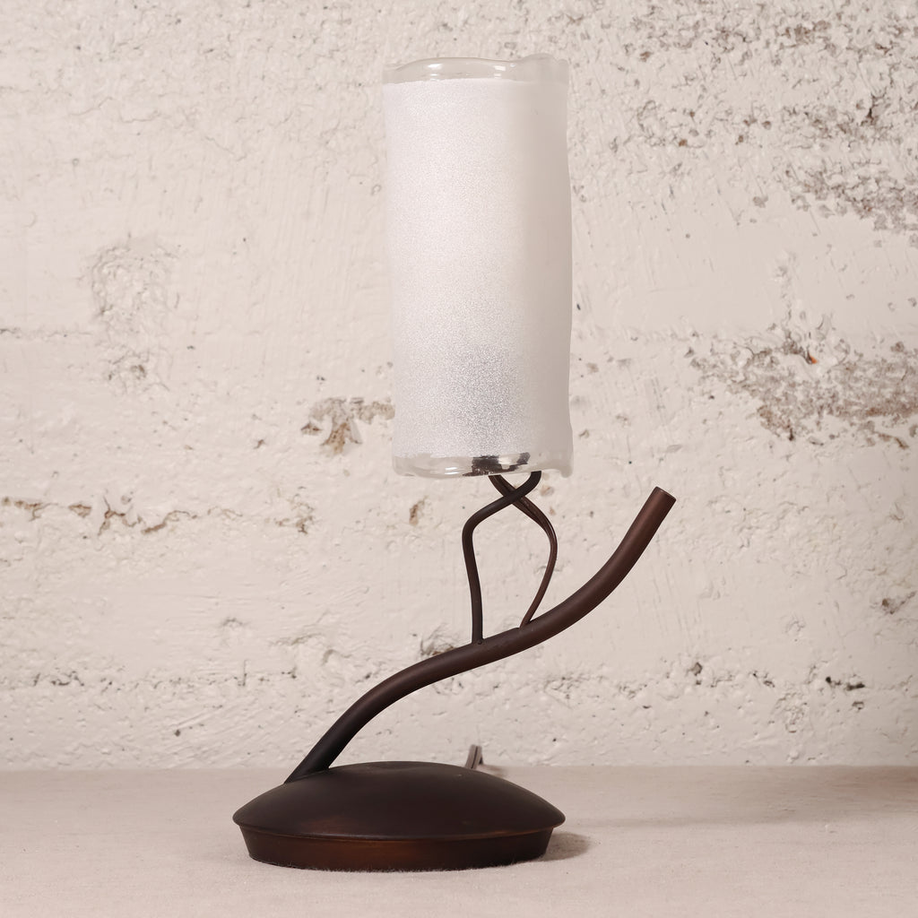 A table lamp with a frosted white cylindrical glass shade and a dark brown sculptural metal base, set against a textured off-white wall.