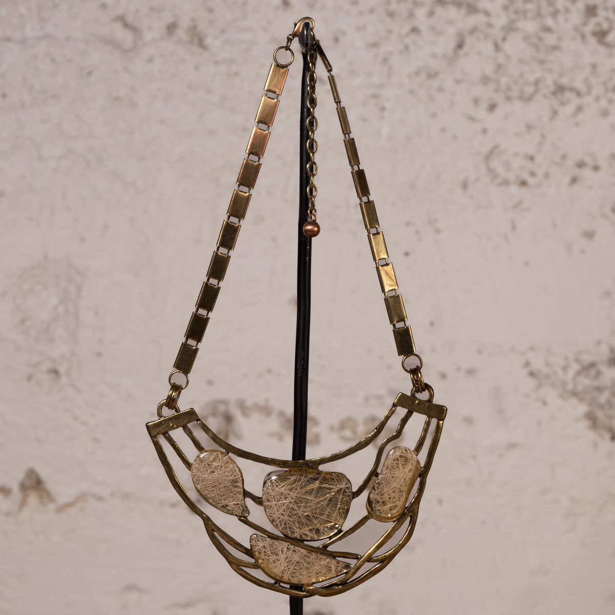 A vintage-style statement necklace with a gold-toned metal frame and five textured, organic-shaped clear resin or glass pendants, hanging on a black display stand against a textured beige wall.