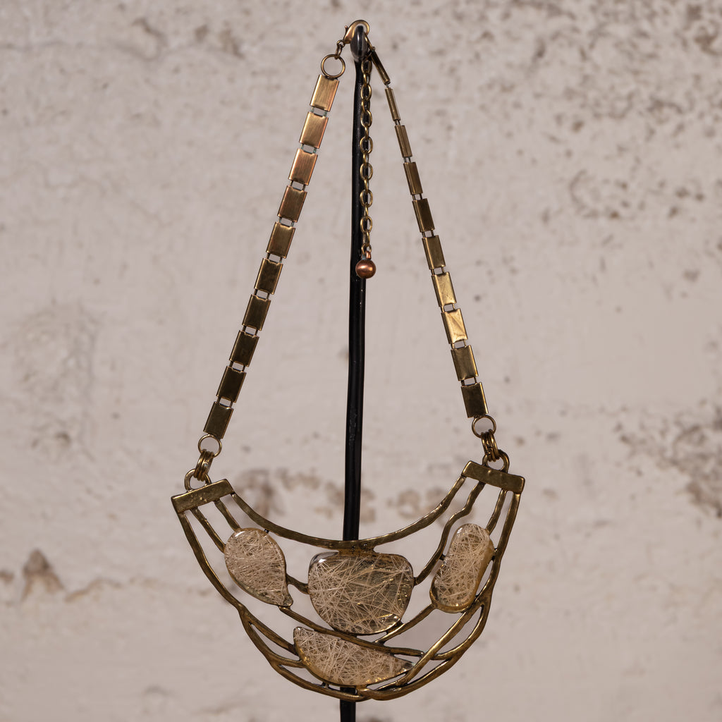 A vintage-style statement necklace with a gold-toned metal frame and five textured, organic-shaped clear resin or glass pendants, hanging on a black display stand against a textured beige wall.