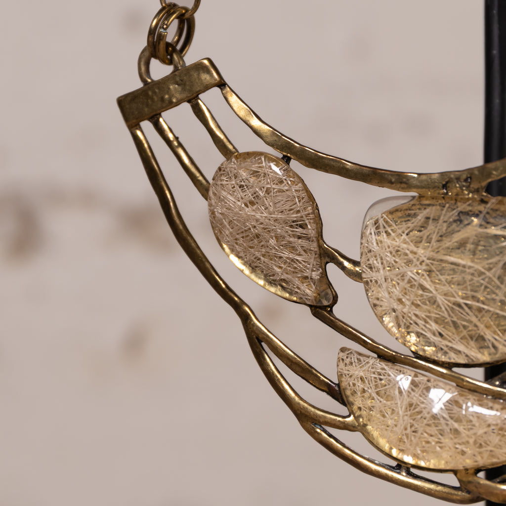Close-up of a crescent-shaped pendant featuring a gold-toned metal frame with three teardrop-shaped clear resin sections filled with fine white fiber, shown against a soft beige background.