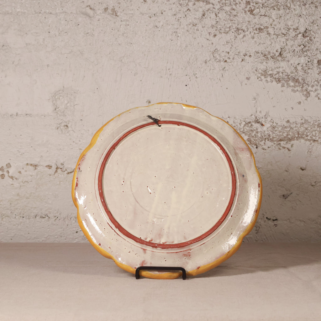 Decorative ceramic plate with a scalloped edge on a stand against a textured wall.