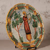 Ceramic plate with a decorative design of a person playing a harp surrounded by trees and fruit on a stand.