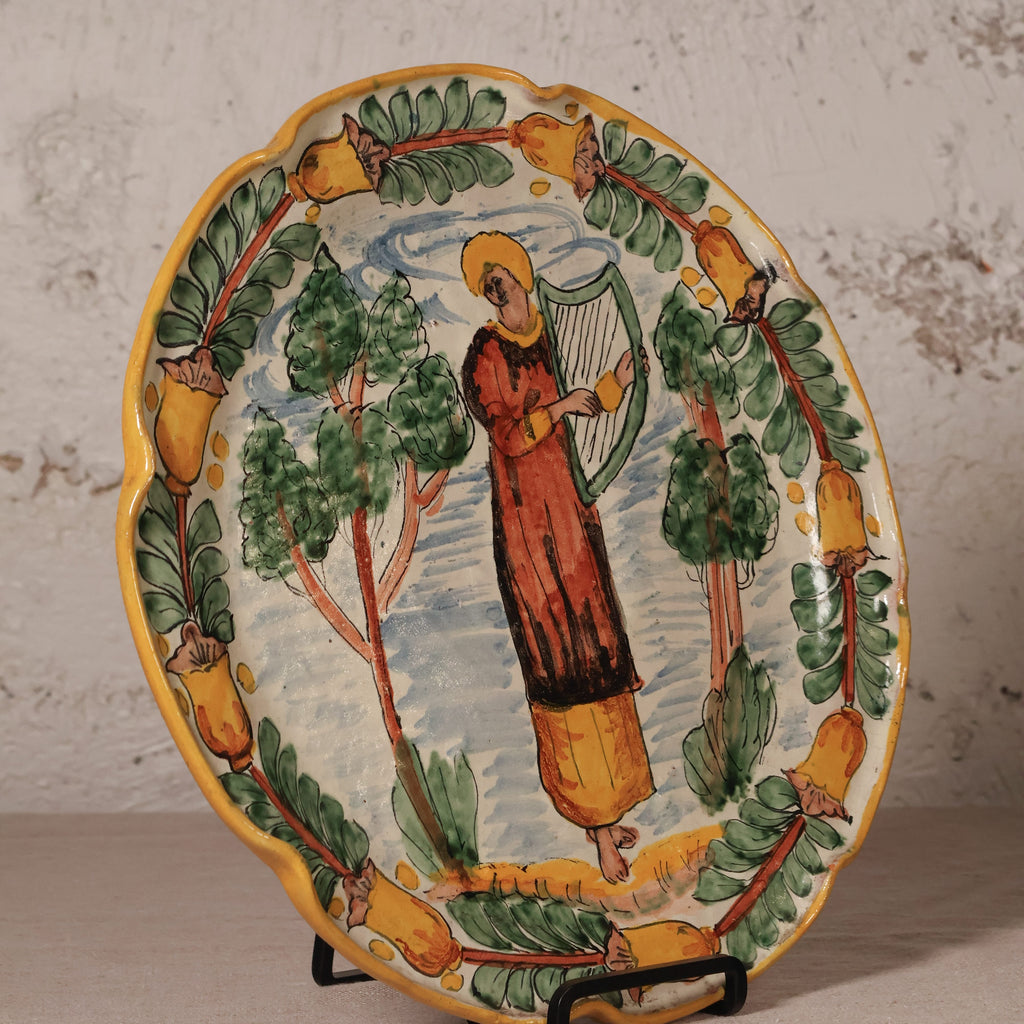 Ceramic plate with a decorative design of a person playing a harp surrounded by trees and fruit on a stand.