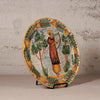 An oval hand-painted ceramic plate displayed on a small black stand against a textured, light beige plaster wall. The plate features a colorful illustration of a figure playing a harp, surrounded by green trees and a border of yellow acorns.