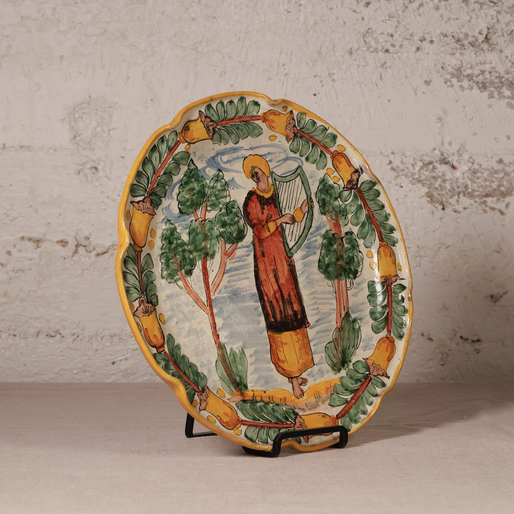 An oval hand-painted ceramic plate displayed on a small black stand against a textured, light beige plaster wall. The plate features a colorful illustration of a figure playing a harp, surrounded by green trees and a border of yellow acorns.