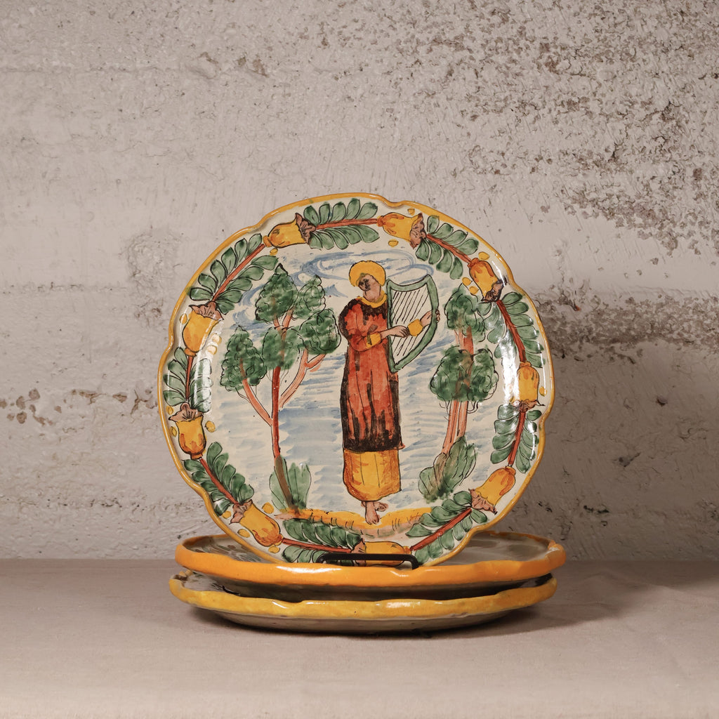 A stack of hand-painted ceramic plates with a rustic Mediterranean style, featuring a central figure playing a harp surrounded by trees and a border of lemons, displayed on a neutral surface against a textured plaster wall.