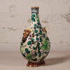 A vintage Italian-style ceramic vase with a bulbous body, narrow neck, and lion head handles, decorated with green floral vines and a cream background against a textured plaster wall.