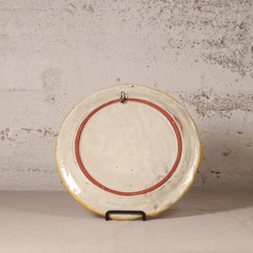 A vintage ceramic plate with a distressed cream glaze, featuring a red painted circular accent and a yellow rim, displayed on a small black stand against a textured plaster wall.