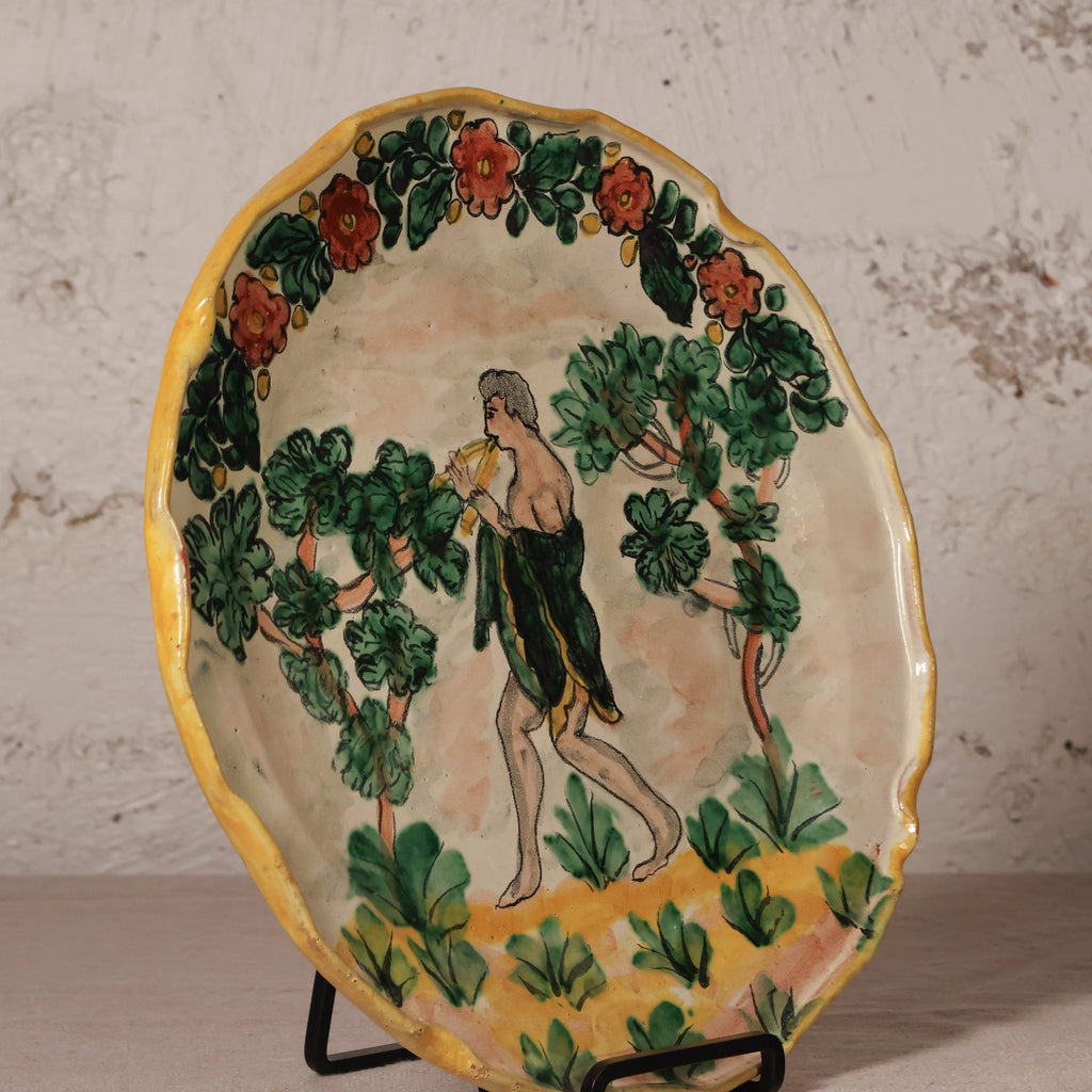 An oval hand-painted ceramic plate with a yellow rim, featuring a classical scene of a nude male figure walking among green trees with red flowers, displayed on a black stand against a textured white wall.