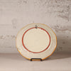 Round ceramic plate with a red rim on a stand against a textured wall.
