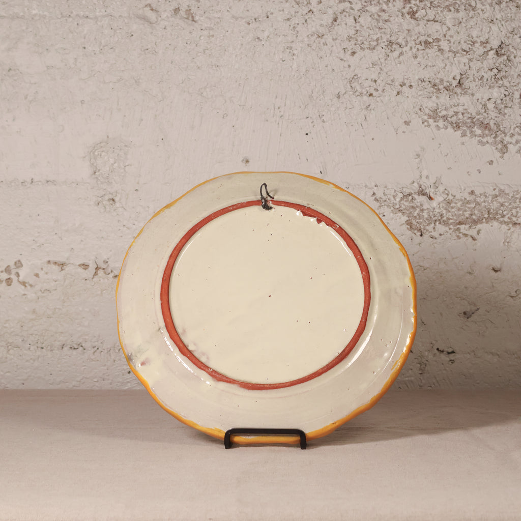 Round ceramic plate with a red rim on a stand against a textured wall.
