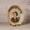 Decorative plate with a painted portrait on a stand against a textured wall.