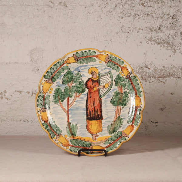 Decorative plate with a person playing a harp and surrounded by trees and birds on a textured wall background.