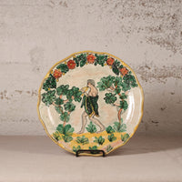 A vintage hand-painted ceramic plate with a fluted rim, featuring a classical figure walking among leafy green trees and orange flowers, displayed on a small stand against a textured light gray wall.