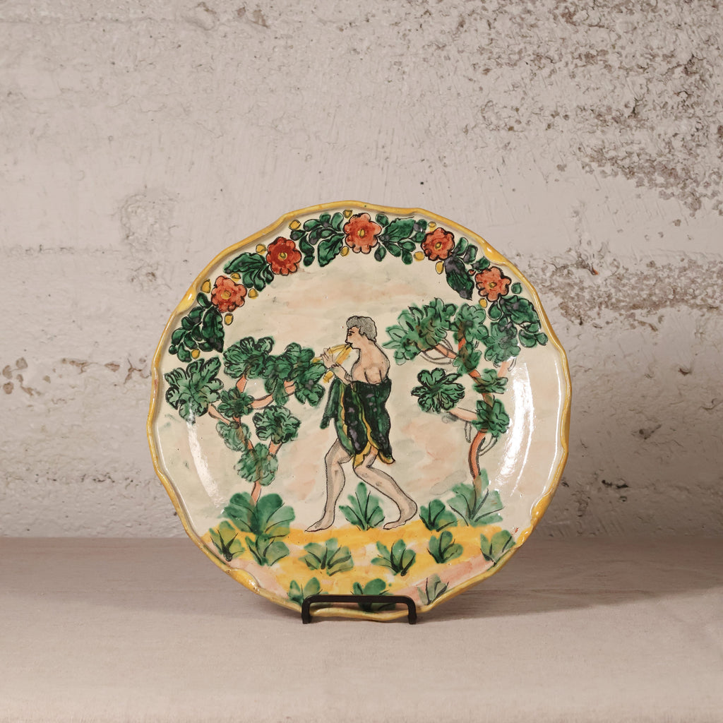 A vintage hand-painted ceramic plate with a fluted rim, featuring a classical figure walking among leafy green trees and orange flowers, displayed on a small stand against a textured light gray wall.