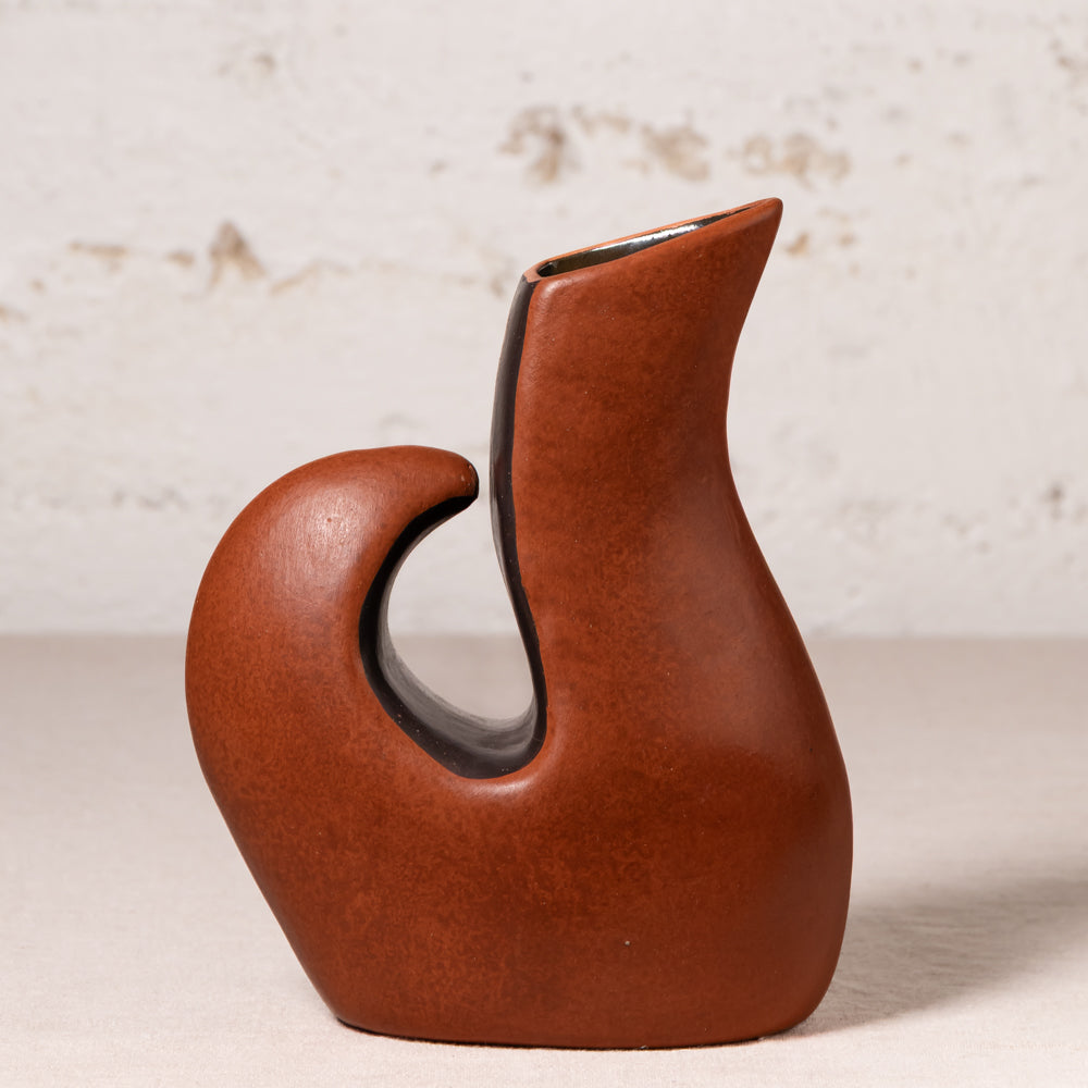 An abstract sculptural ceramic vase with a curved handle and a narrow neck, finished in a warm reddish-brown glaze with a dark interior, set against a light textured wall.