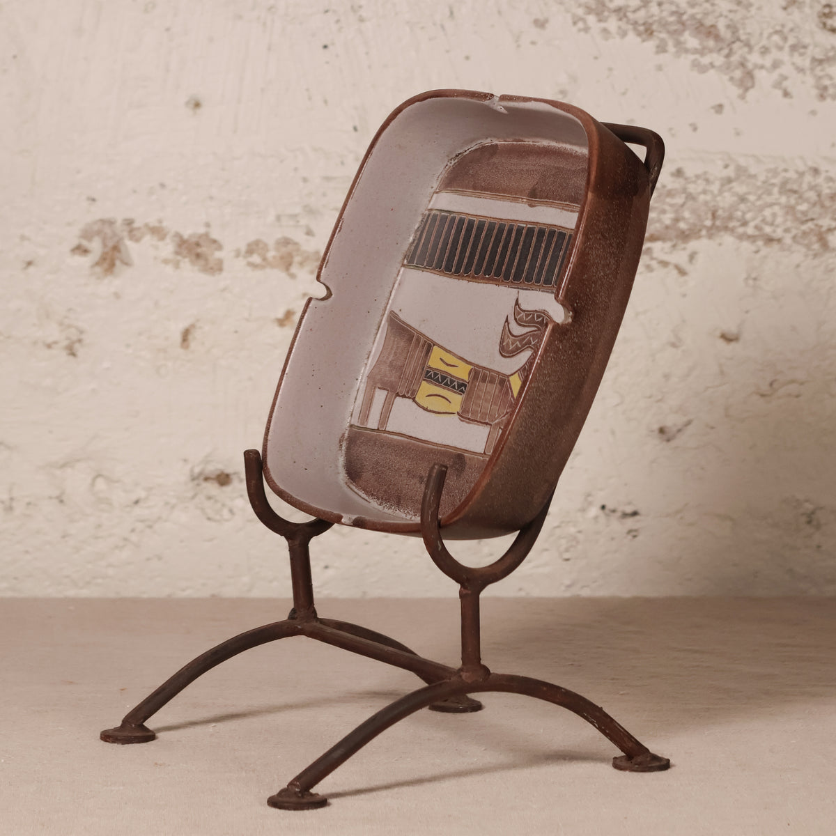 A decorative ceramic ashtray featuring a vintage car illustration, displayed on a rusted metal stand against a textured, distressed off-white wall.