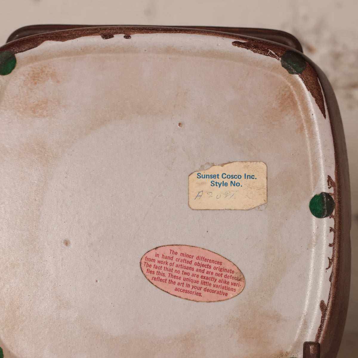 The underside of a square ceramic decorative object, showing a cream-colored glazed interior, a brown rim, small green tabs at the corners, and two labels: one reading 'Sunset Cosco Inc. Style No.