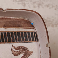 Close-up of a handmade ceramic tray with a rustic finish, featuring a brown stylized animal motif and a black-and-brown striped band, set against a textured beige background.
