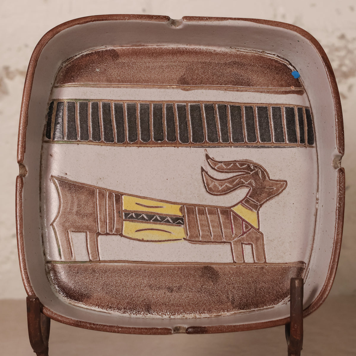 A square ceramic ashtray with rounded corners, featuring a stylized Egyptian cartouche design with a dog figure in brown, black, and yellow on a beige background.
