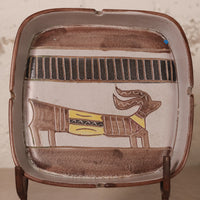 A square ceramic ashtray with rounded corners, featuring a stylized Egyptian cartouche design with a dog figure in brown, black, and yellow on a beige background.