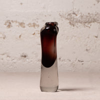 A small hand-blown glass bottle with a dark amber-brown top section and a clear glass bottom, standing upright against a textured off-white wall.