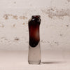 A small hand-blown glass bottle with a dark amber-brown top section and a clear glass bottom, standing upright against a textured off-white wall.