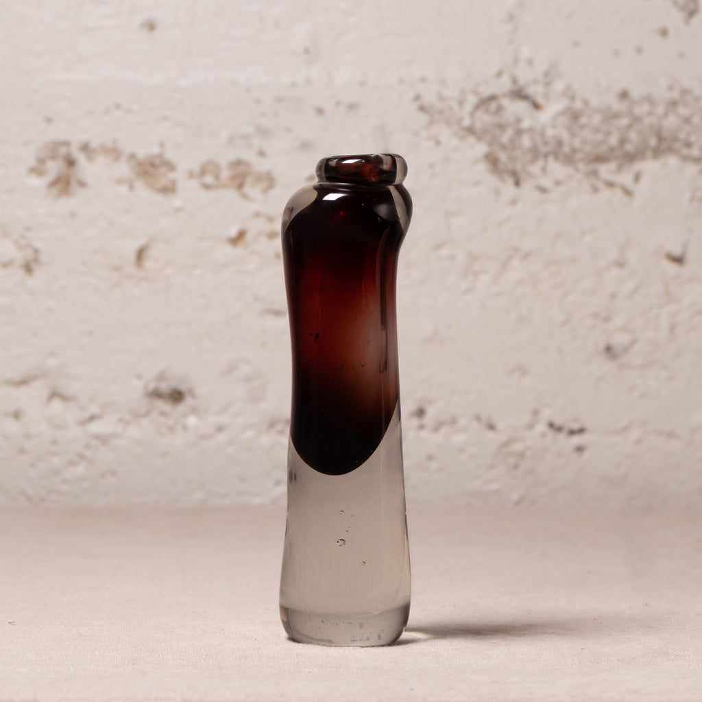 A small hand-blown glass bottle with a dark amber-brown top section and a clear glass bottom, standing upright against a textured off-white wall.