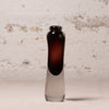 A small hand-blown glass bottle with a dark amber-brown bulbous neck and a clear glass base, standing on a light surface against a textured off-white brick wall.