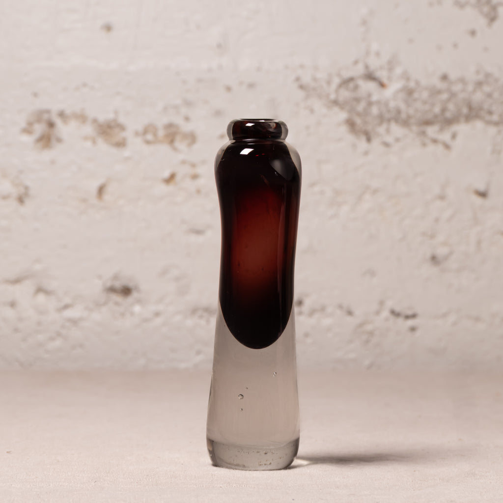 A small hand-blown glass bottle with a dark amber-brown bulbous neck and a clear glass base, standing on a light surface against a textured off-white brick wall.