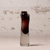 A small hand-blown glass vase with a dark amber-brown top and a translucent smoky gray base, standing on a light surface against a textured off-white wall.