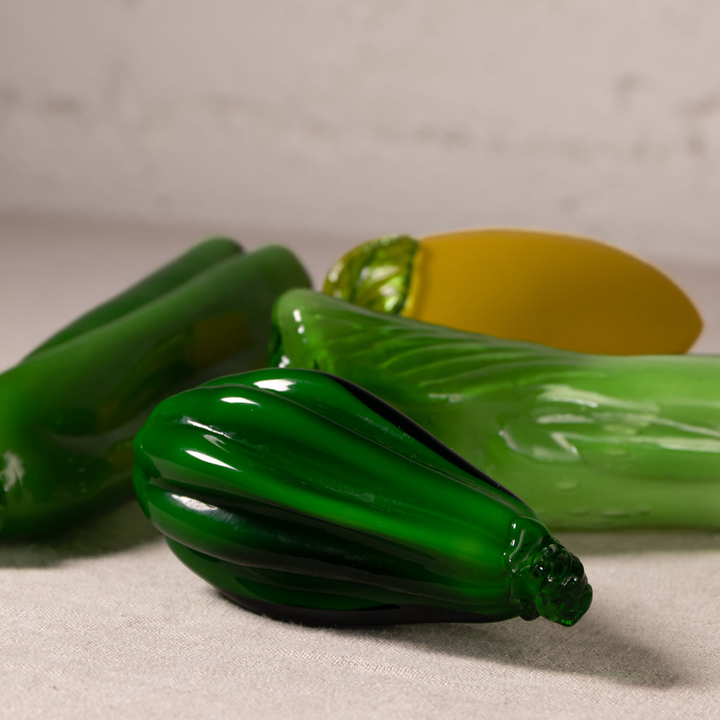 Three glossy glass vegetables, including green zucchini, yellow squash, and green bell peppers, arranged on a neutral textured surface.