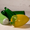 Three translucent glass vegetable figurines, including a yellow pepper, green zucchini, and a light green cucumber, arranged on a textured beige surface against a light gray background.