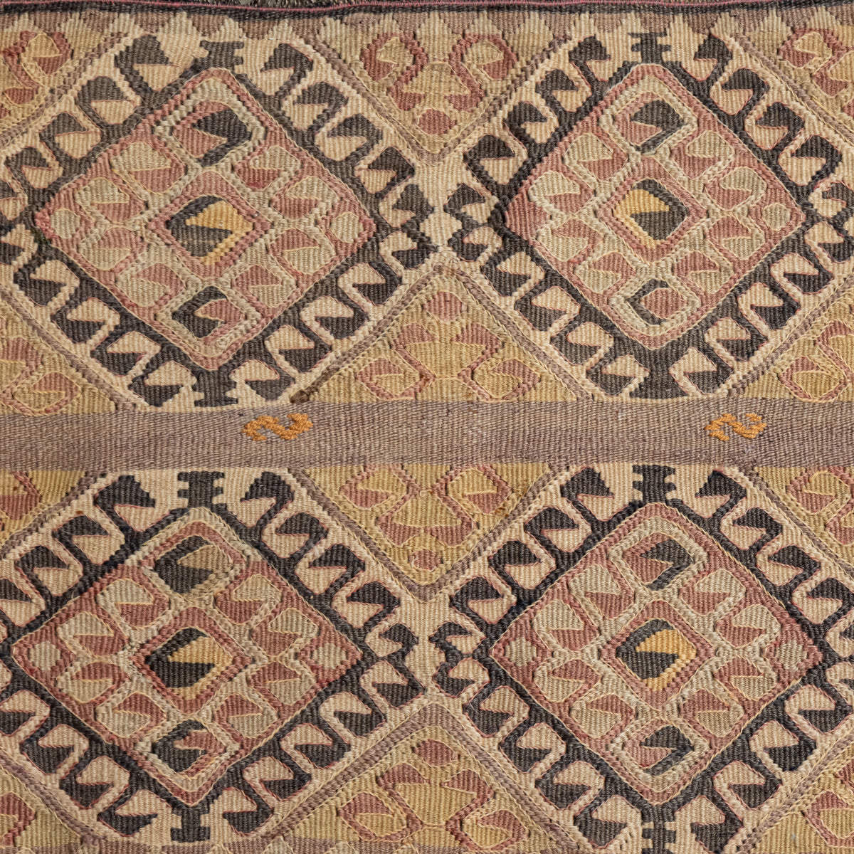 A close-up of a woven textile with a repeating geometric diamond pattern in beige, brown, muted red, and black, with a horizontal band of solid tan woven through the center.