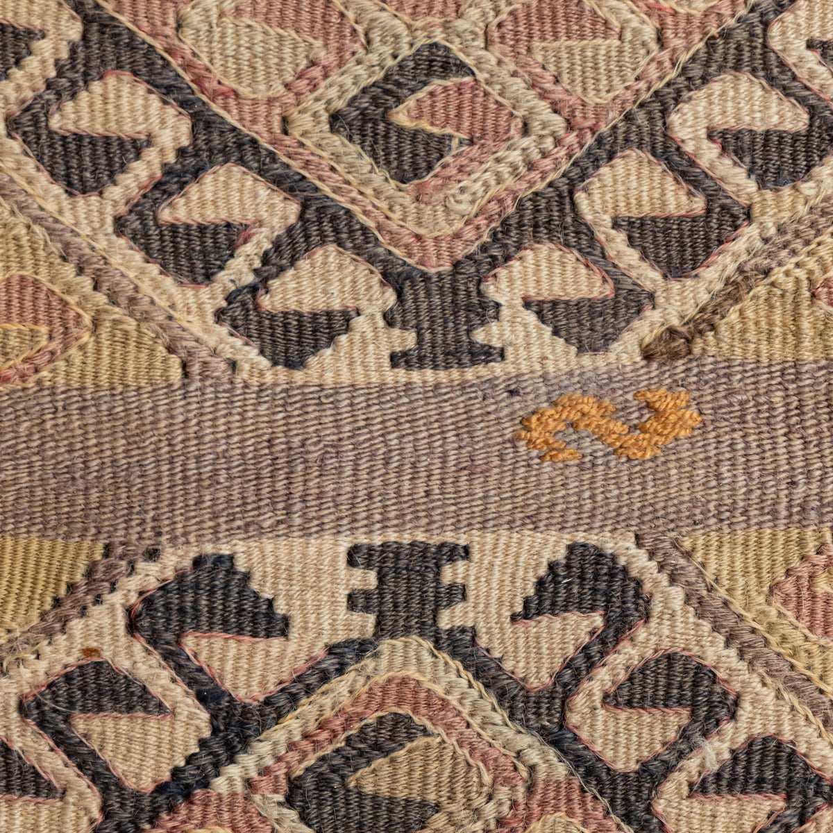 Close-up of a woven textile with a geometric pattern in beige, brown, muted pink, and charcoal, plus a horizontal band with a small orange motif.
