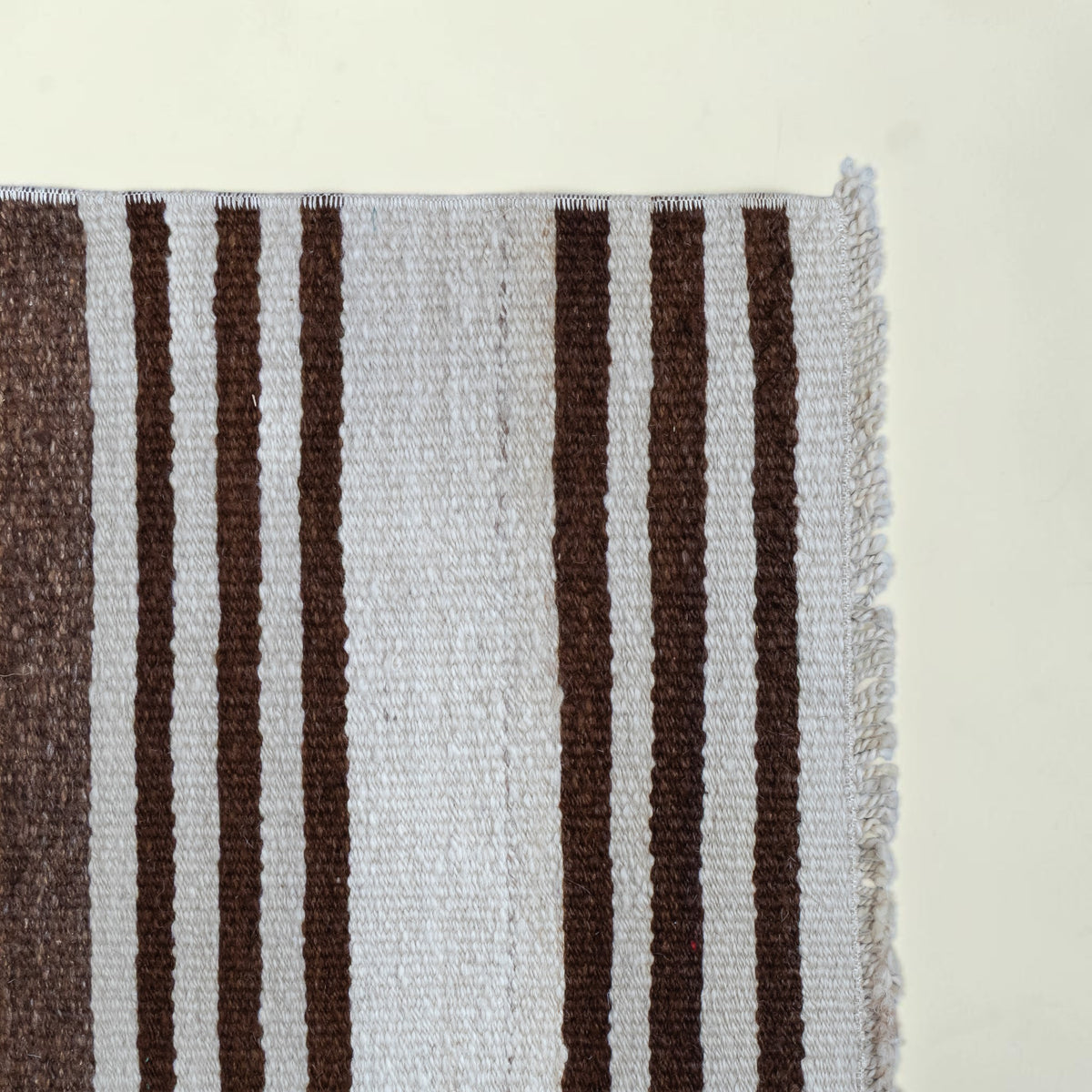 A close-up of a woven rug corner showing vertical stripes in dark brown and off-white, with a frayed edge on the right side.