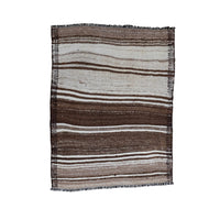 A rectangular woven placemat with vertical stripes in brown, beige, and white, featuring fringed edges, displayed flat against a plain off-white background.