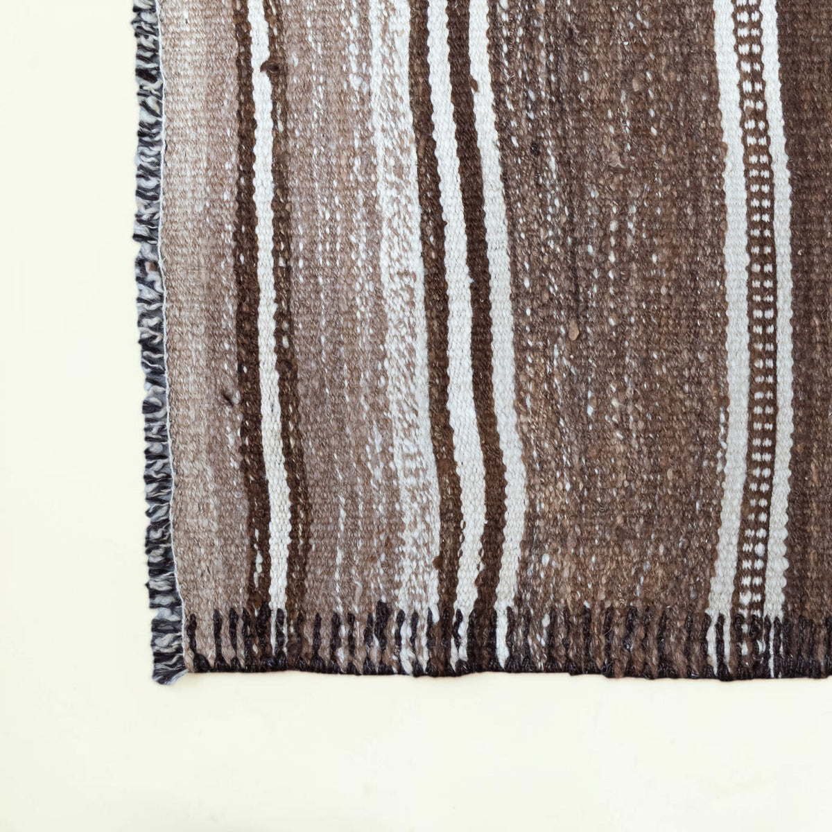 Close-up of a woven textile with vertical brown, taupe, and off-white stripes and dark fringe along one edge, shown against a plain light background.
