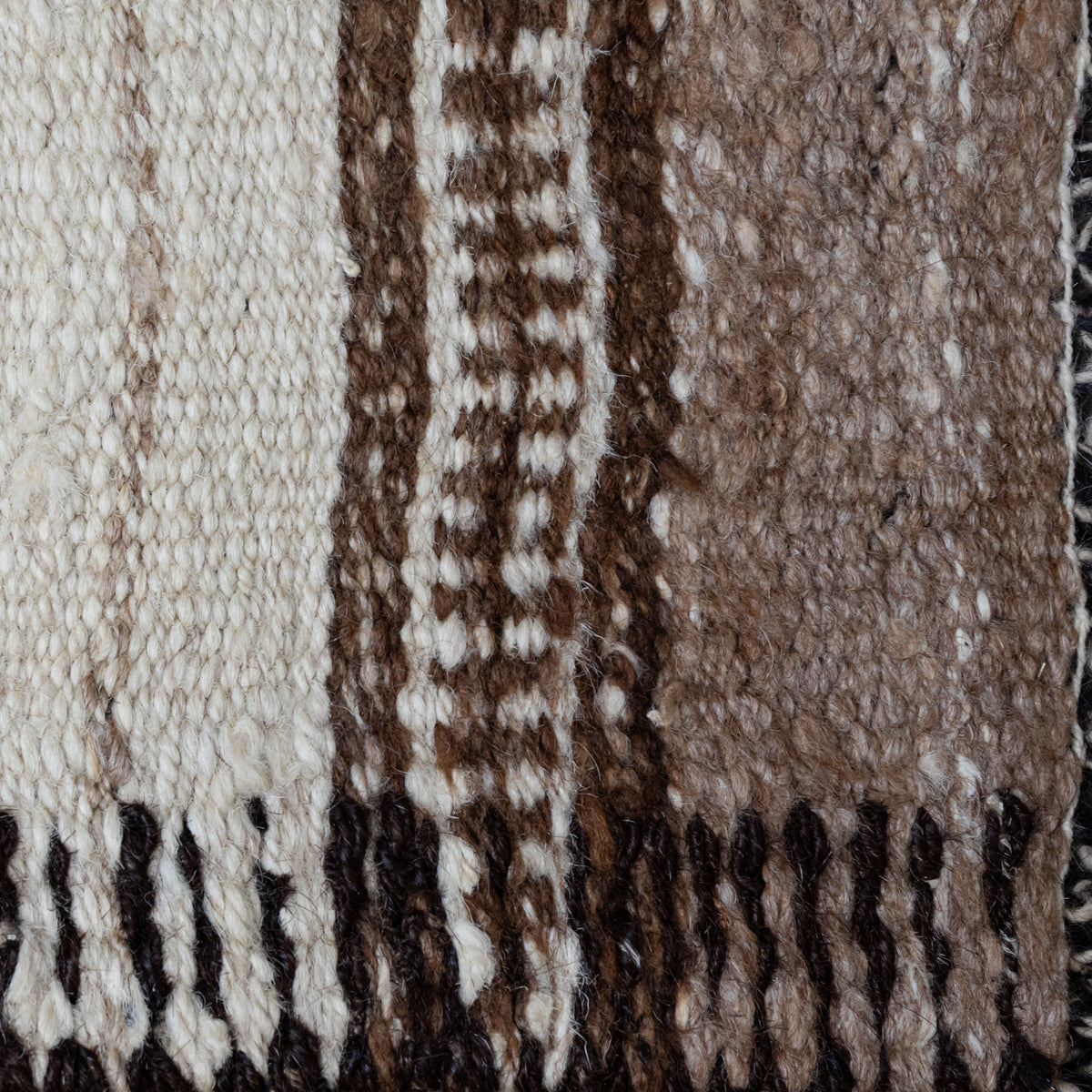 Close-up of a woven textile with vertical stripes in cream, brown, and taupe, finished with dark fringe at the bottom.