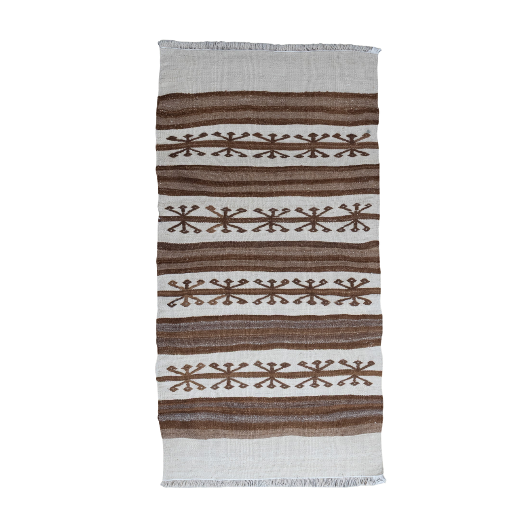 A rectangular woven textile with fringe on the top and bottom edges, featuring horizontal stripes in brown and off-white with geometric snowflake-like motifs, shown against a plain white background.