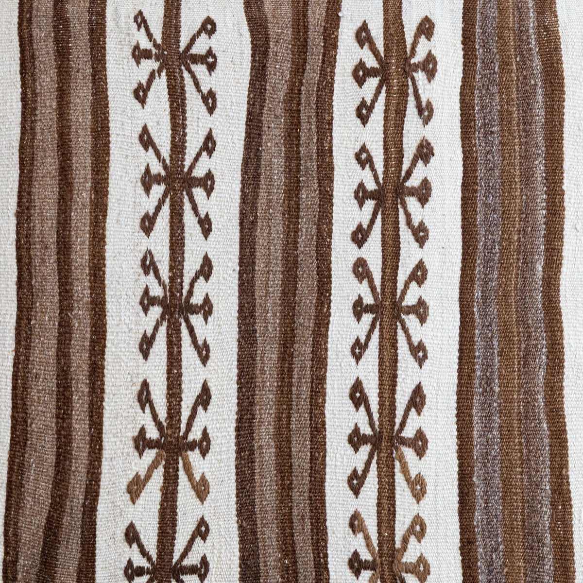 A close-up of a woven textile with vertical stripes in brown and off-white, featuring repeating geometric diamond and cross-like motifs.