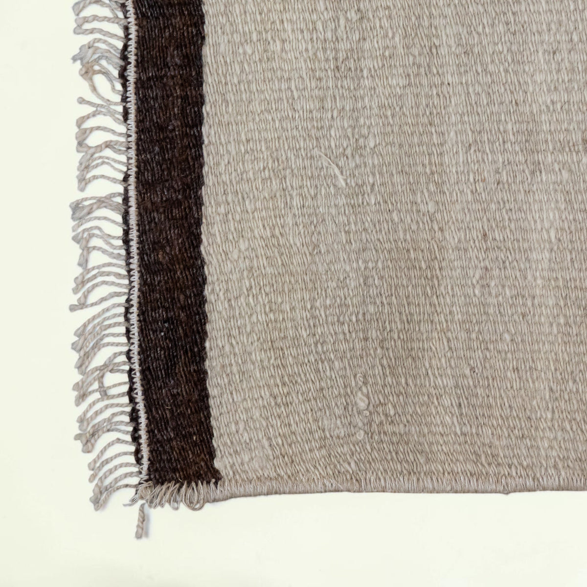 Close-up of a woven rug corner showing a light beige main field with a dark brown vertical stripe and fringed edge on a white background.