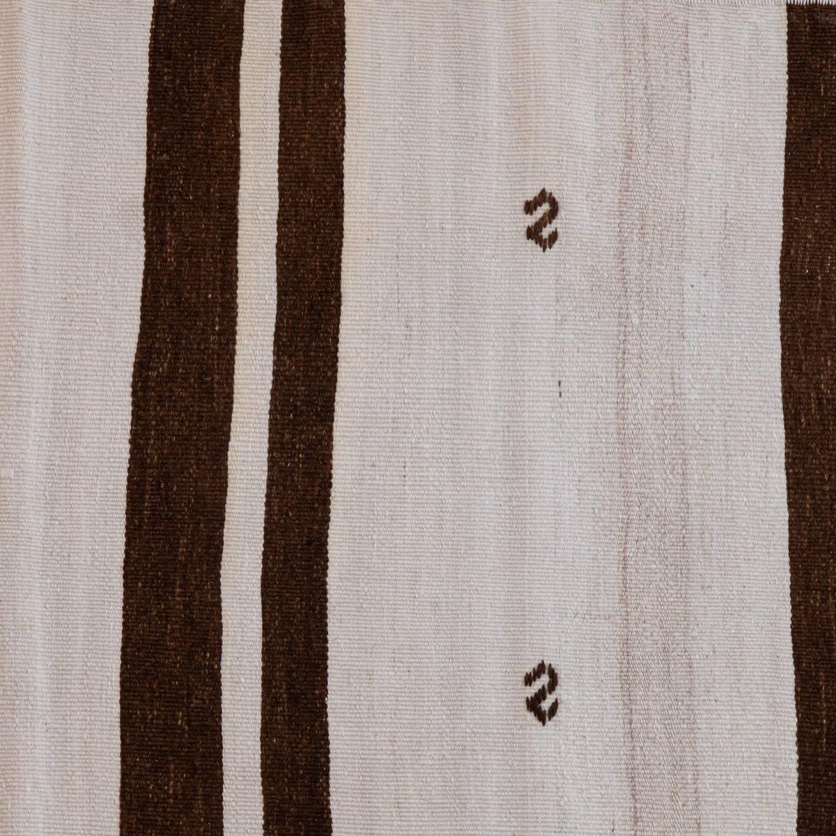 Close-up of a woven textile with a cream background, two wide dark brown vertical stripes, and two small dark brown zigzag motifs on the right side.
